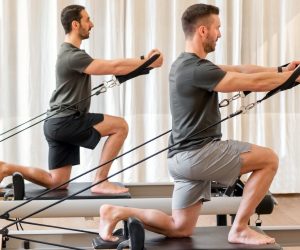 Full body side view of strong men in sportswear doing hug tree kneeling pilates exercise with resistance bands on reformer bed during training in gym